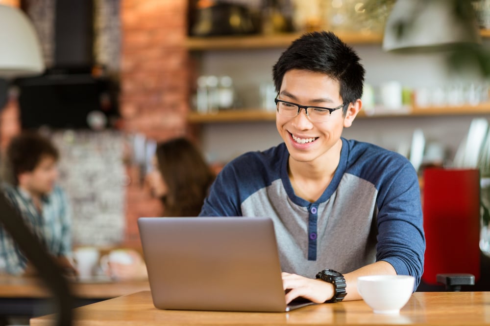 Happy cheerful young asian male in glasses smiling and using laptop in cafe-1